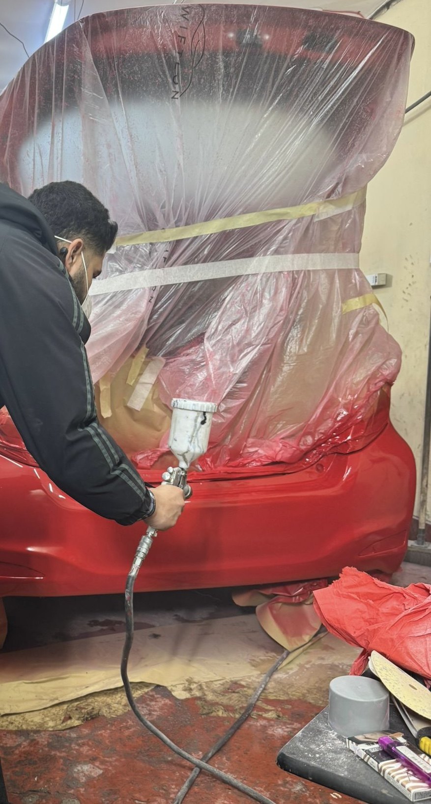 A sprayer painting the rear of a red car that has been wrapped in masking film